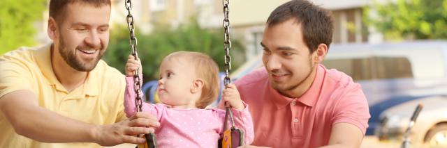 Zwei Männer spielen mit ihrer Tochter, die auf einer Schaukel sitzt.