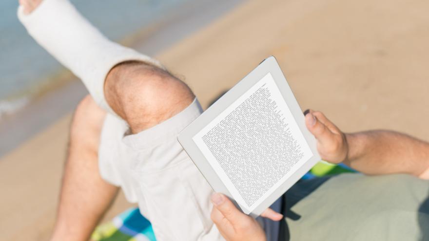 Jemand liegt mit einem Bein im Gips am Strand und liest ein Buch.