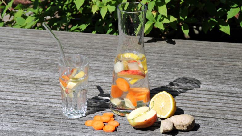 Glas und Karaffe mit Wasser und Stücken von Möhren, Äpfeln, Zitronen und Ingwer