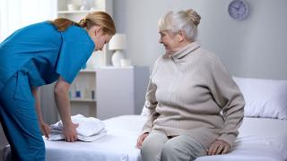 Eine ältere Dame sitz auf ihrem Bett, eine Pflegerin legt Bettwäsche neben ihr ab