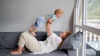 Frau oiegt auf Sofa und hebt Kleinkind vor Fensterfront hoch