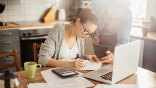 Eine Frau sitzt mit Laptop, Taschenrechner, Rechnungen und Stift am Küchentisch, ein Mann steht neben ihr und beugt sich auf eine Rechnung blickend zu ihr herunter.